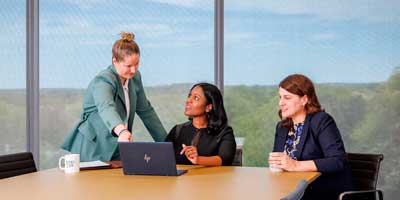 Three professional women in a business meeting, collaborating at a conference table with a laptop in an office with large windows and a scenic view in the background.