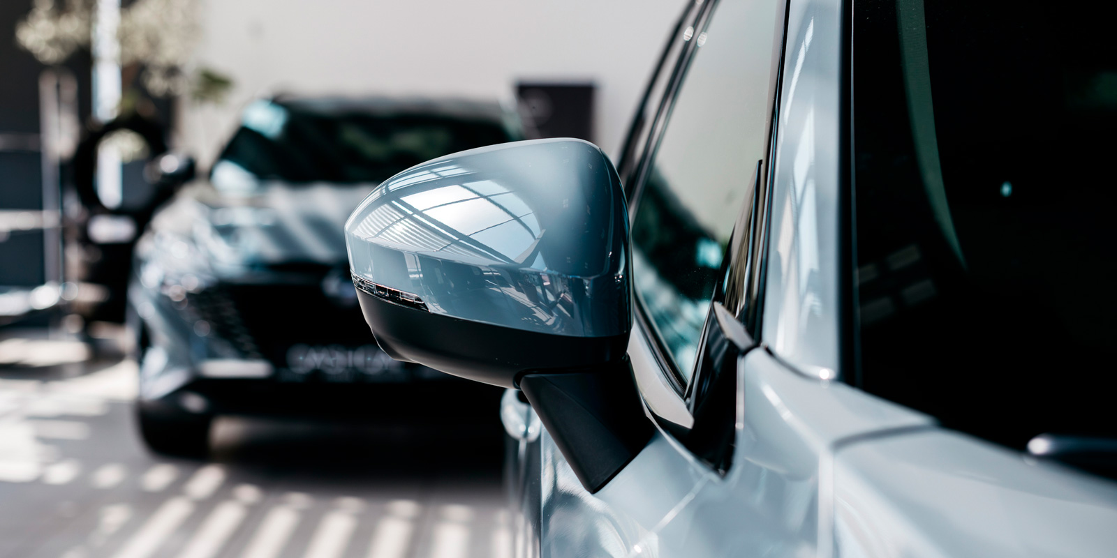 Close-up of a modern car side mirror with another vehicle blurred in the background at a dealership.