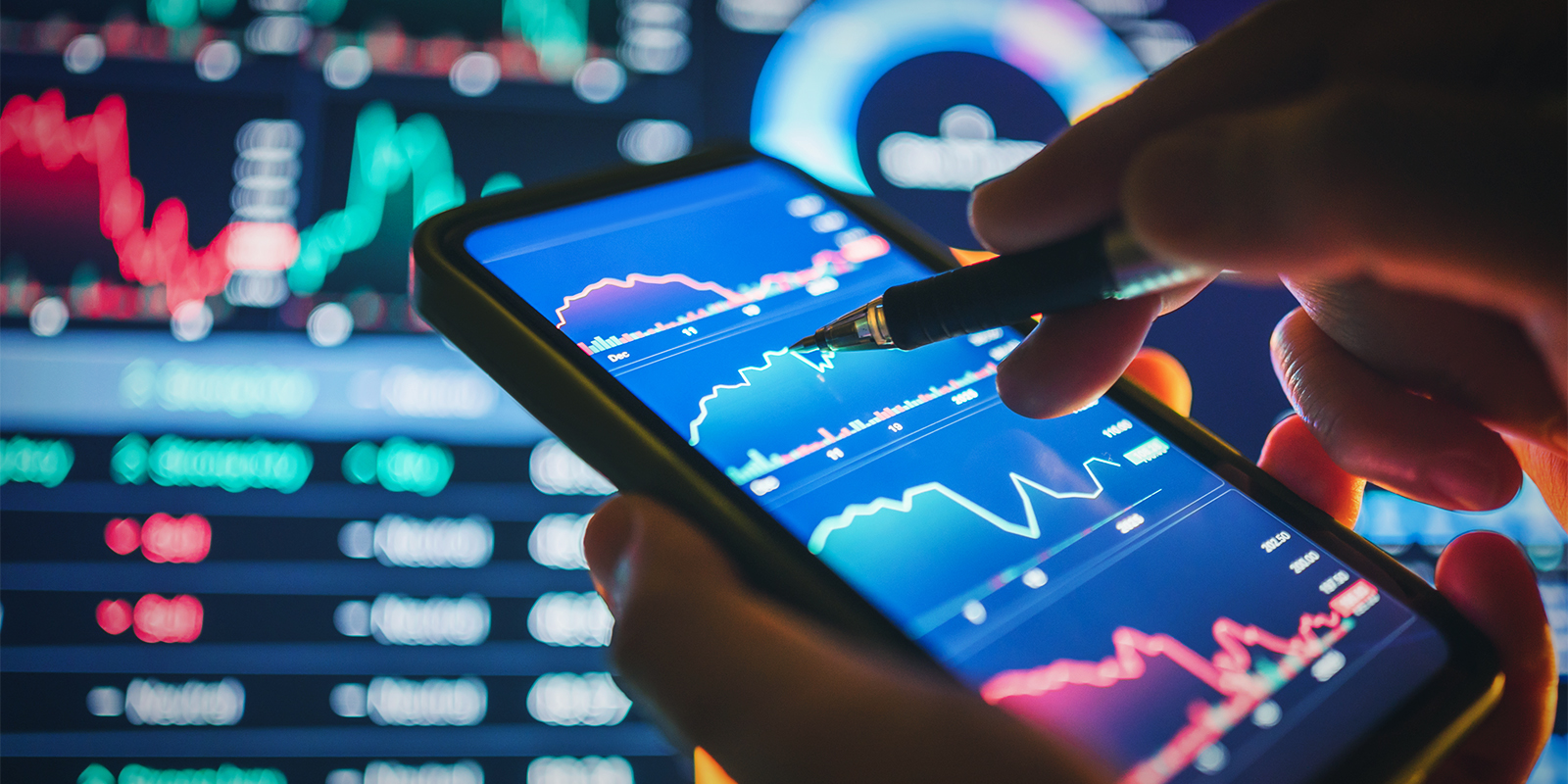 Close-up of a person analyzing financial charts on a smartphone with stock market data and graphs displayed on screens in the background, symbolizing mobile trading and investment analysis.